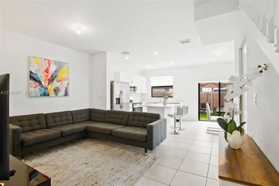 Open-concept interior featuring light-toned tile flooring, recessed ceiling lighting, a kitchen island with stone-surface countertop, and a sliding glass door providing natural light