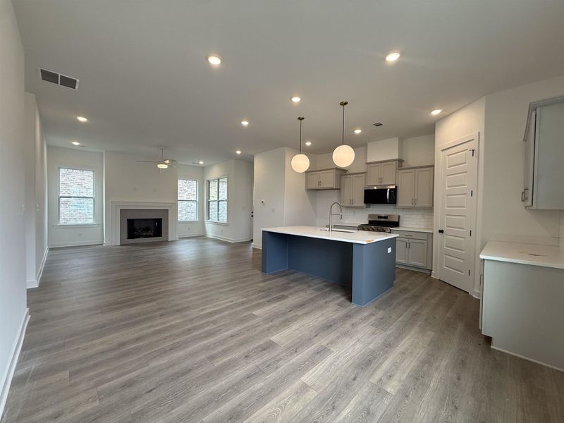 Open-concept interior featuring wood-finish flooring throughout, a central kitchen island with pendant lighting, recessed ceiling lighting, a fireplace with a mantel, and multiple windows