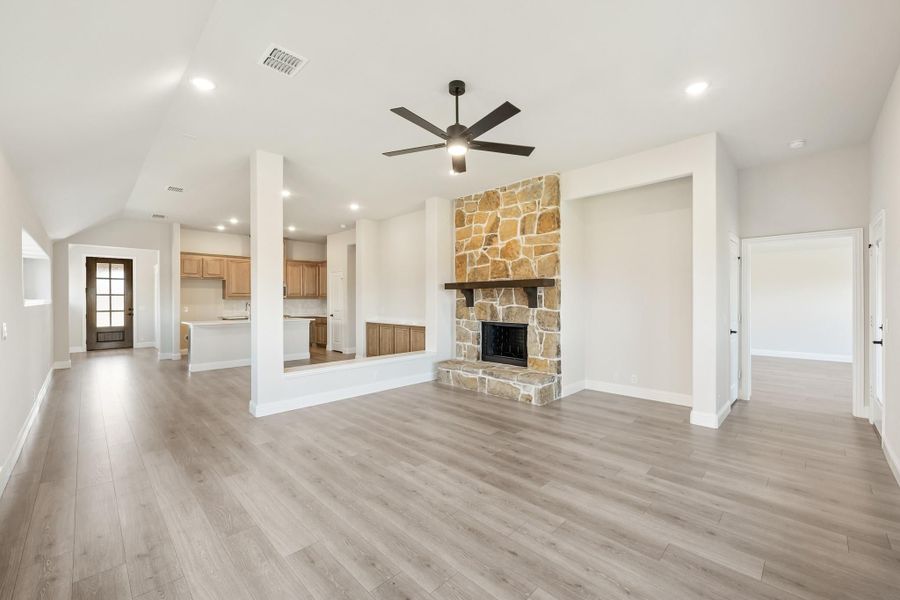 Open living room with stone fireplace, hardwood floors, ceiling fan, and open kitchen in background Open living room with stone fireplace, hardwood floors, ceiling fan, and open kitchen in background