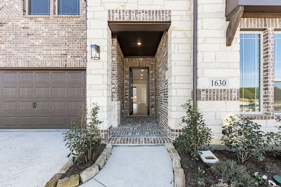 Covered front entry with brick and stone exterior, walkway, and attached garage at home 1630 Covered front entry with brick and stone exterior, walkway, and attached garage at home 1630
