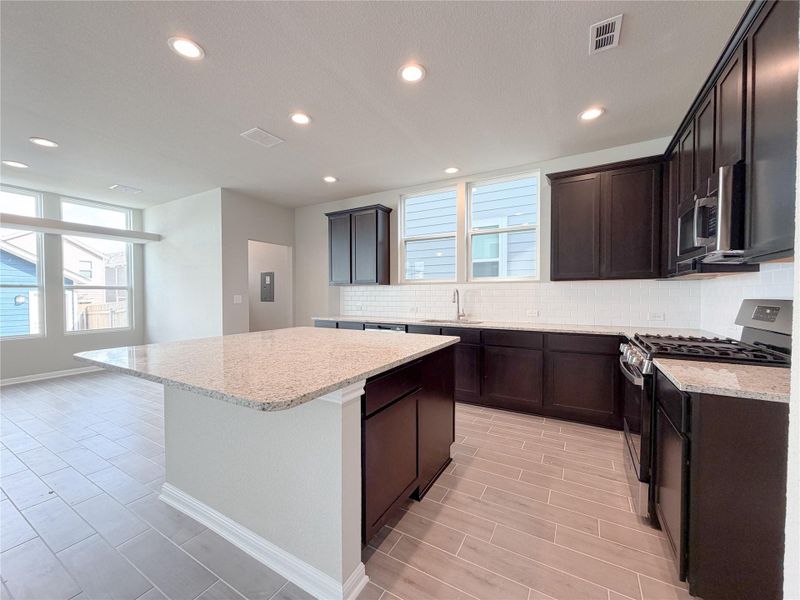 Contemporary kitchen featuring an expansive island with a light-toned countertop, dark wood-finish cabinetry, subway tile backsplash, stainless steel appliances, and recessed lighting