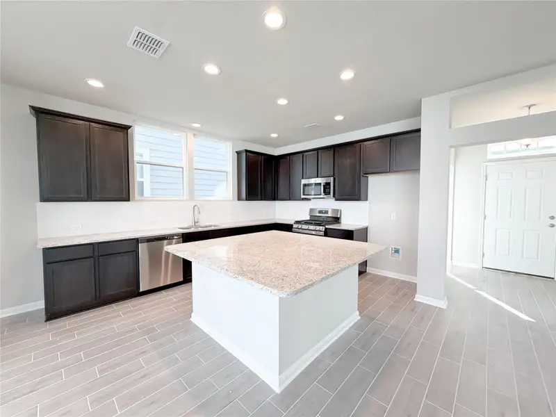 Modern kitchen featuring a large central island with a light-toned countertop, dark wood-finish cabinetry, stainless steel appliances, a white subway tile backsplash, and wood-look tile flooring