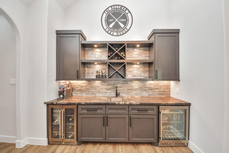 Elegant wet bar featuring dark cabinetry, stone backsplash, built-in wine storage, and dual glass-door beverage coolers, perfect for entertaining.