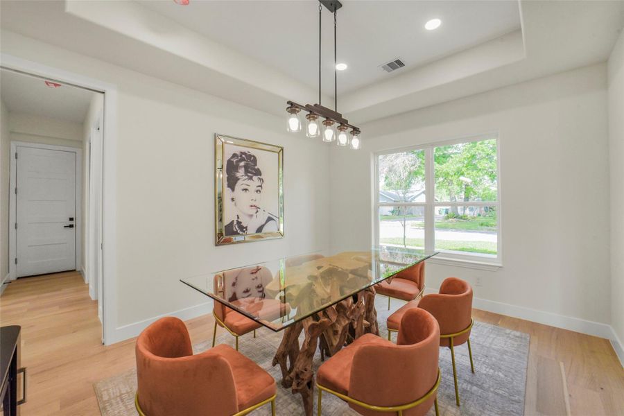 This dining area features a unique glass-top table with a natural wood base, surrounded by stylish orange chairs. Large windows provide ample natural light, complemented by recessed ceiling lighting. A modern artwork adds a chic touch to the neutral-toned walls.
