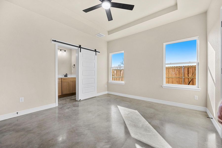 Unfurnished bedroom featuring a barn door, concrete floors, a ceiling fan, ensuite bathroom, and a tray ceiling