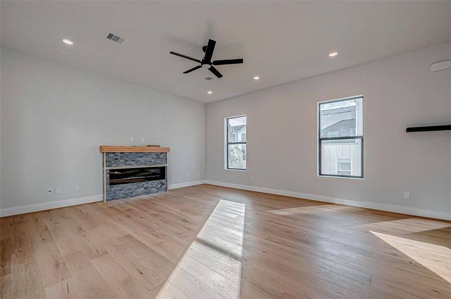 Bright, spacious living room with hardwood floors, modern ceiling fan, and a stylish stone fireplace. Large windows provide ample natural light, creating a welcoming atmosphere.