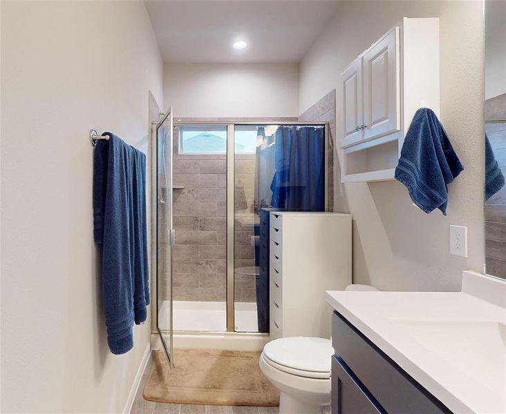 This bathroom features a modern design with a glass-enclosed shower, sleek gray tile, a white vanity with a large mirror, and ample storage including a wall-mounted cupboard.