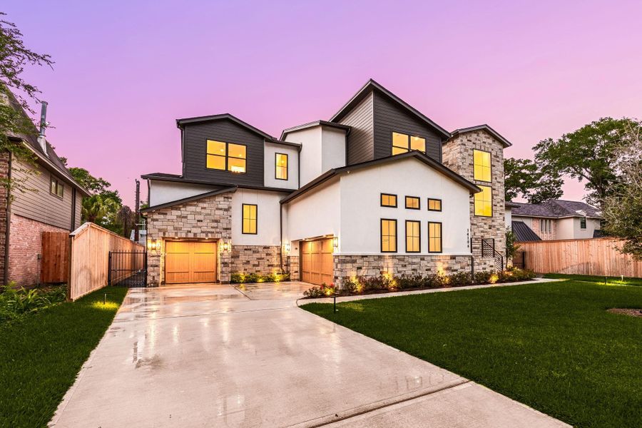 A striking first impression with clean architectural lines, smooth stucco, and layered stonework brought to life under a glowing evening sky. Warm exterior lighting highlights the textures and thoughtful design, while expansive windows hint at the light-filled interiors within. The wide driveway and dual garages add everyday functionality to the home’s bold, modern presence, creating a refined yet welcoming approach from the moment you arrive.
