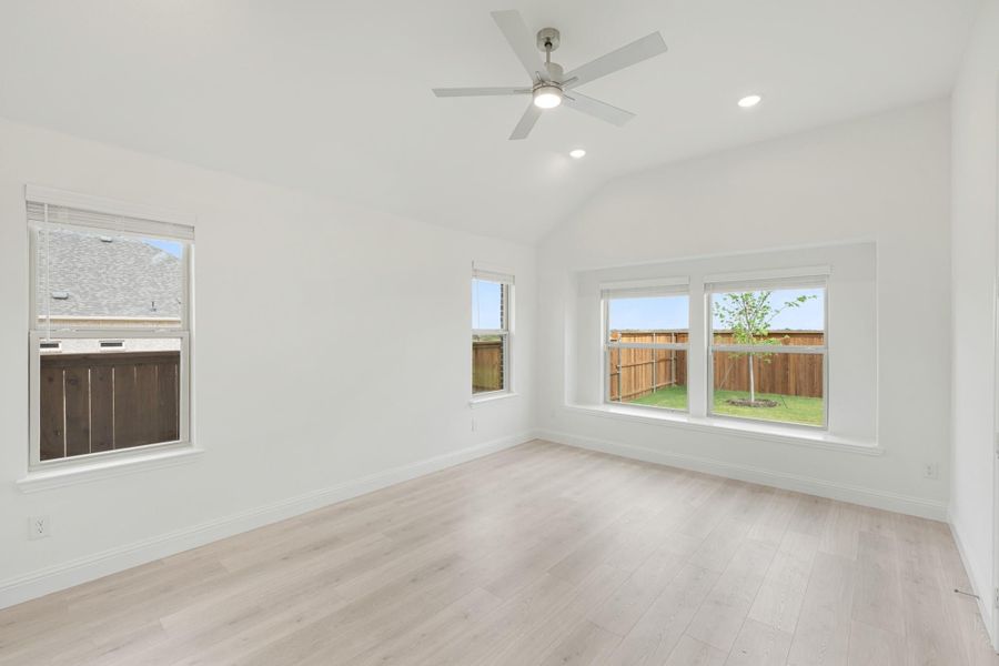 Empty room with vaulted ceiling, ceiling fan, light wood floors, and multiple windows overlooking fenced backyard