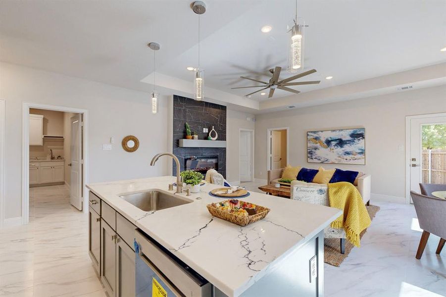 Kitchen featuring a center island with sink, a tiled fireplace, ceiling fan, a tray ceiling, and sink