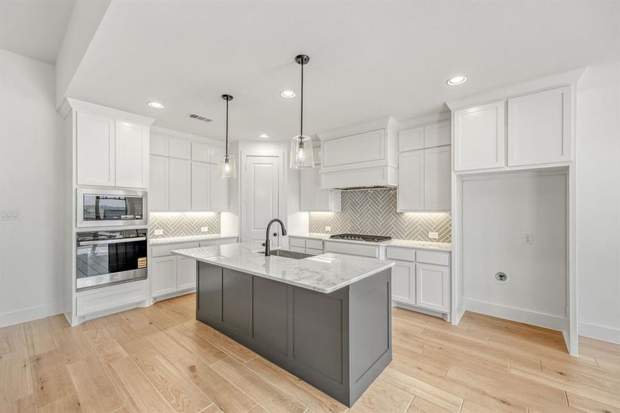 Kitchen featuring two tone cabinets, stainless steel appliances, an island with sink, hanging light fixtures, and backsplash