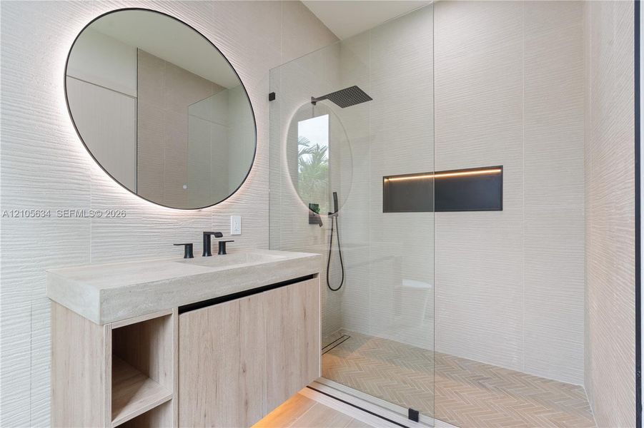 Floating vanity with integrated sink, textured wall tile, circular backlit mirror, frameless glass shower enclosure with rainfall showerhead, and herringbone-patterned shower floor tile
