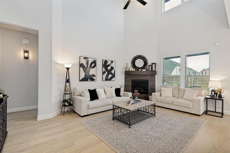 Two-story living area featuring a stacked stone fireplace with a wood mantel, high ceilings, large windows, wood-finish flooring, and a ceiling fan