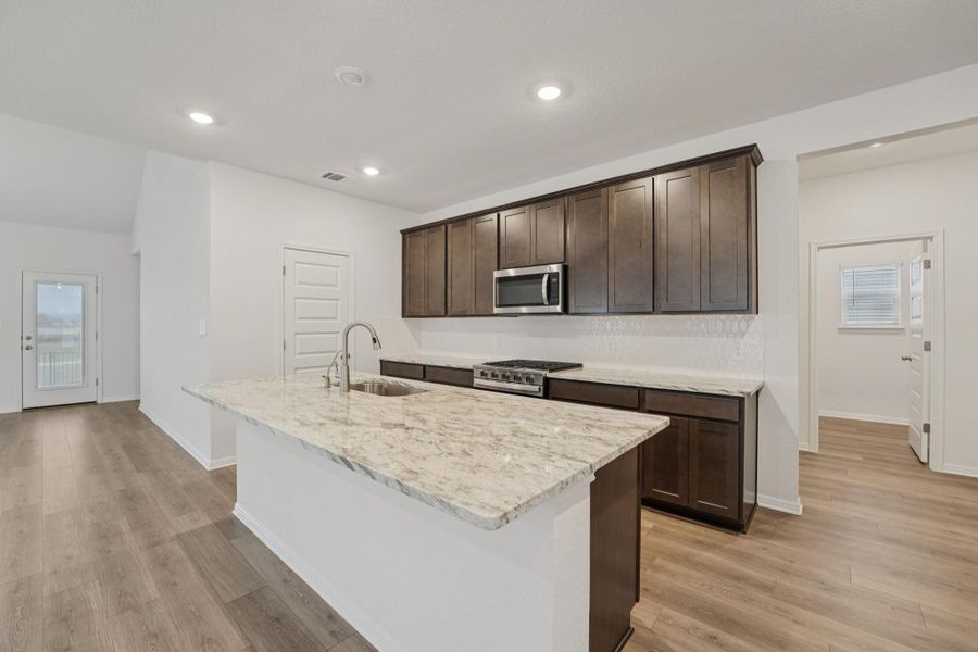 Kitchen with dark brown cabinetry, an island with sink, stainless steel appliances, light stone countertops, and light wood-type flooring