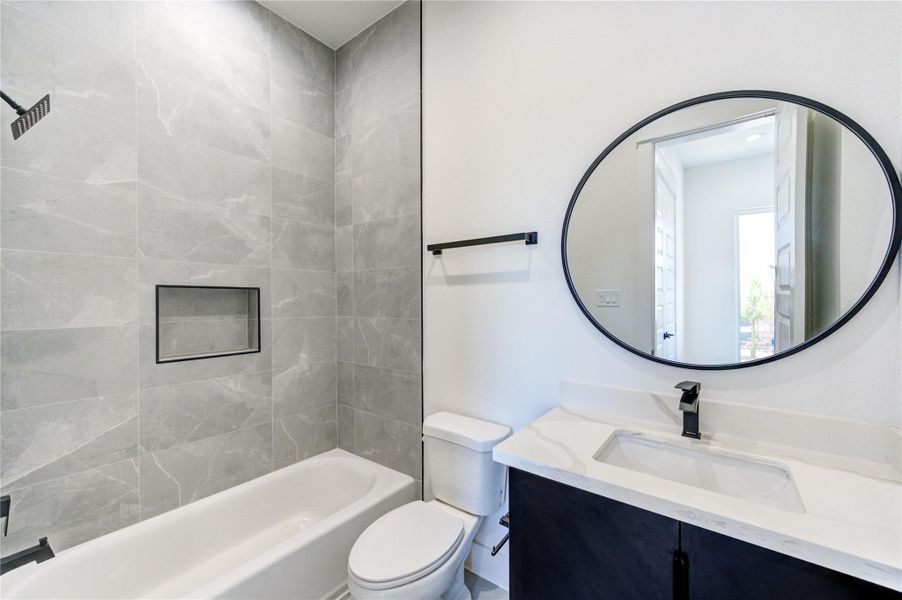 This modern bathroom features sleek floor to ceiling tiles, a bathtub with a built-in niche, and a stylish vanity with a round mirror. It offers a clean, contemporary look with black fixtures and ample natural light.