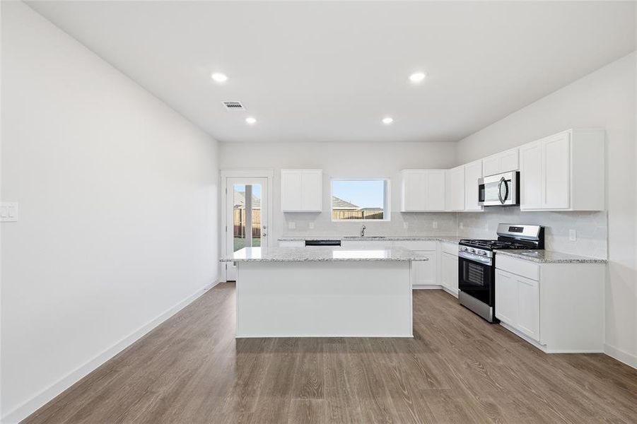 Open-concept kitchen featuring an island, white cabinetry, integrated stainless steel appliances, a tile backsplash, and light-toned countertops Open-concept kitchen featuring an island, white cabinetry, integrated stainless steel appliances, a tile backsplash, and light-toned countertops