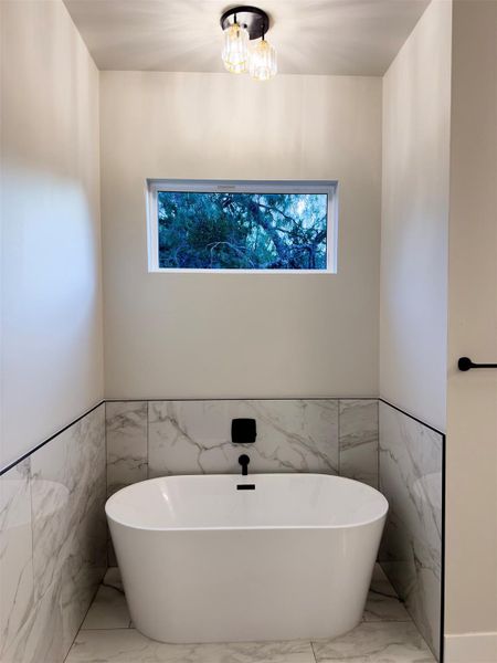 An elegant soaking tub stands positioned against marble-patterned tile underneath a high horizontal window. An elegant soaking tub stands positioned against marble-patterned tile underneath a high horizontal window.