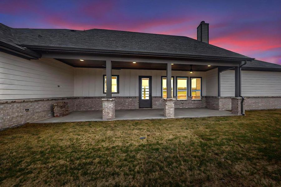 Back of property at dusk featuring roof with shingles, board and batten siding, a patio area, a yard, and a chimney Back of property at dusk featuring roof with shingles, board and batten siding, a patio area, a yard, and a chimney
