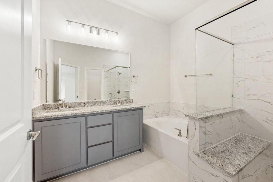 Bathroom with gray double vanity, granite countertops, soaking tub, and glass shower with marble tile walls