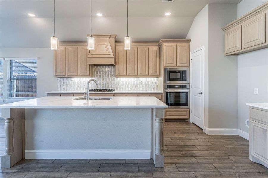 Spacious kitchen featuring a large island with a countertop, wood-finish flooring, built-in stainless steel appliances, a decorative range hood, and pendant lighting