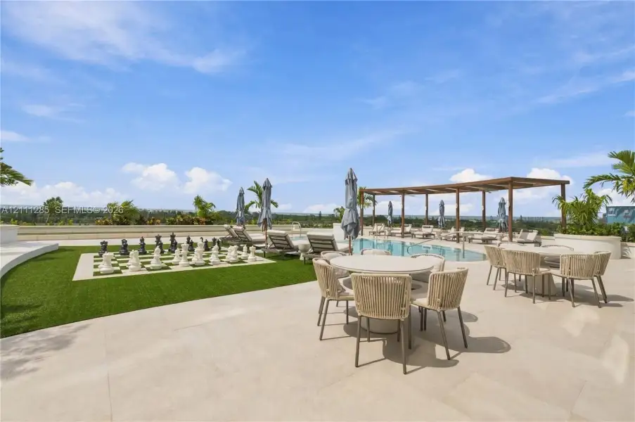 Outdoor terrace featuring light-toned stone paving, a swimming pool, a wood-frame pergola, synthetic turf, and an oversized permanent outdoor chess board Outdoor terrace featuring light-toned stone paving, a swimming pool, a wood-frame pergola, synthetic turf, and an oversized permanent outdoor chess board