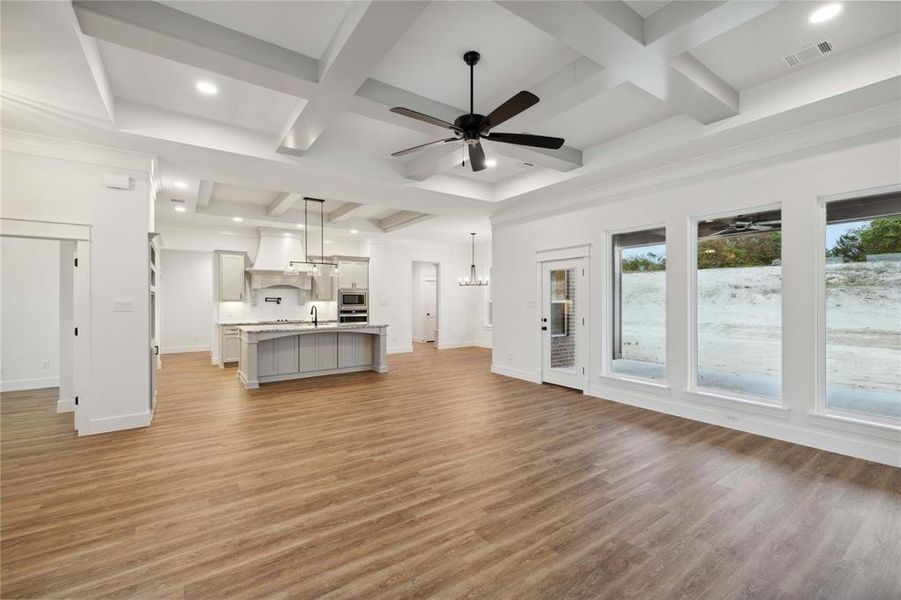 Unfurnished living room with plenty of natural light, ceiling fan with notable chandelier, wood-type flooring, and coffered ceiling