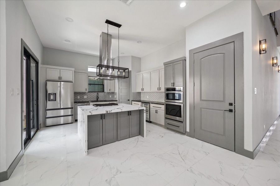 The kitchen showcases a striking island clad in marble-style surfaces, creating a strong focal point for the space. Soft gray cabinetry is paired with a polished tile backsplash, delivering a refined and timeless look.