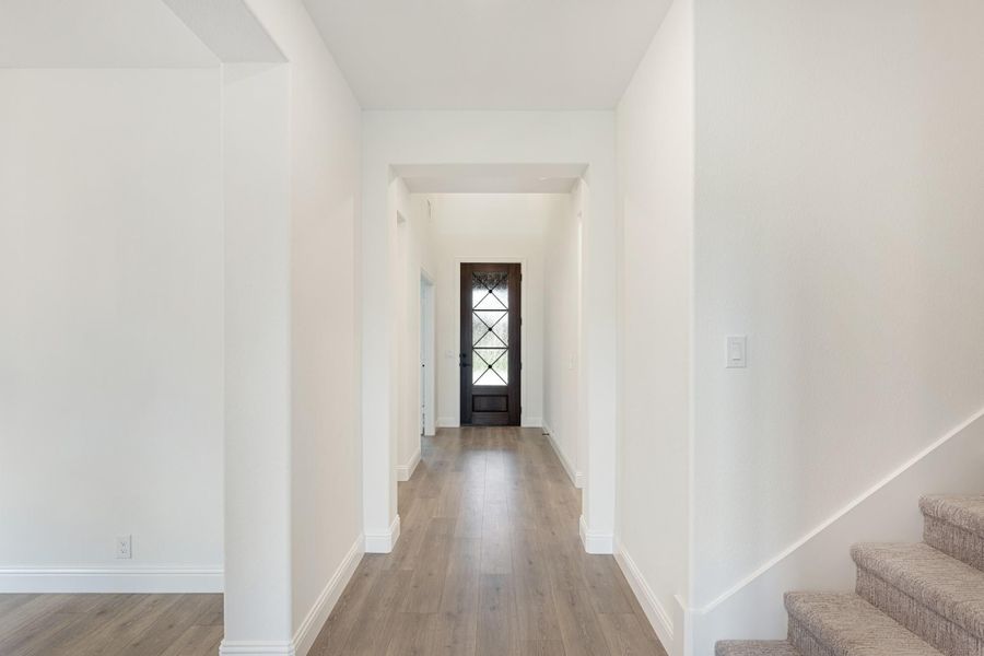 Bright entryway hallway with wood floors, white walls, dark front door with glass panels, and staircase on right. Bright entryway hallway with wood floors, white walls, dark front door with glass panels, and staircase on right.