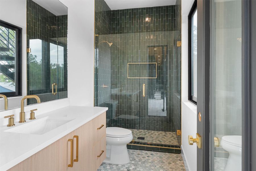 Bathroom featuring a light wood vanity with gold hardware, a white countertop with an integrated sink, a large mirror, and a glass-enclosed shower with dark green subway tiles and a hexagonal tiled floor