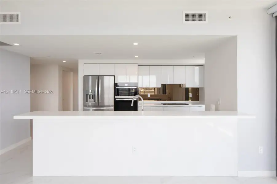 Modern kitchen featuring high-gloss white cabinetry, expansive stone-finish peninsula, stainless steel french-door refrigerator, integrated double oven, and mirrored tile backsplash