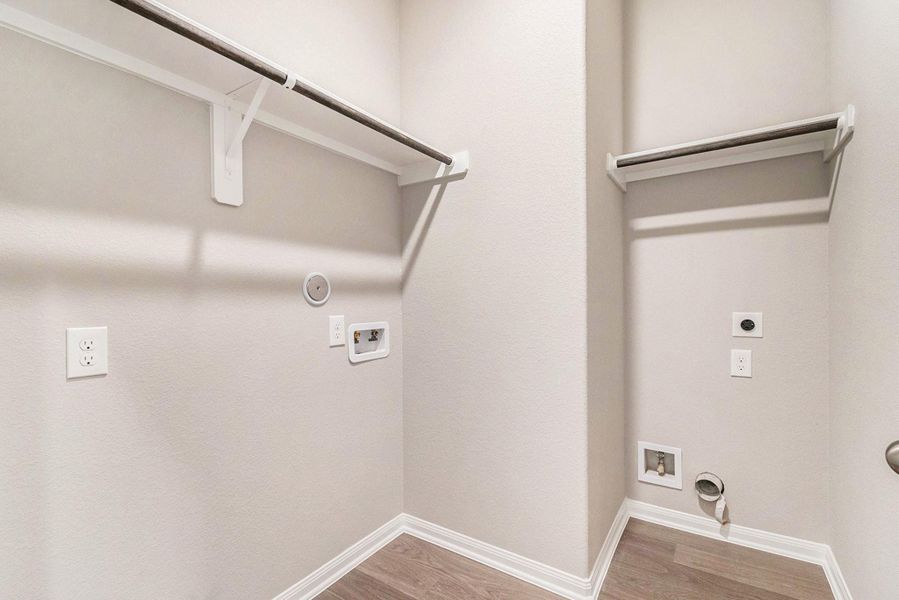 This photo shows a spacious laundry room with hookups for a washer and dryer, featuring light-colored walls and wooden flooring, along with two hanging rods for additional storage.