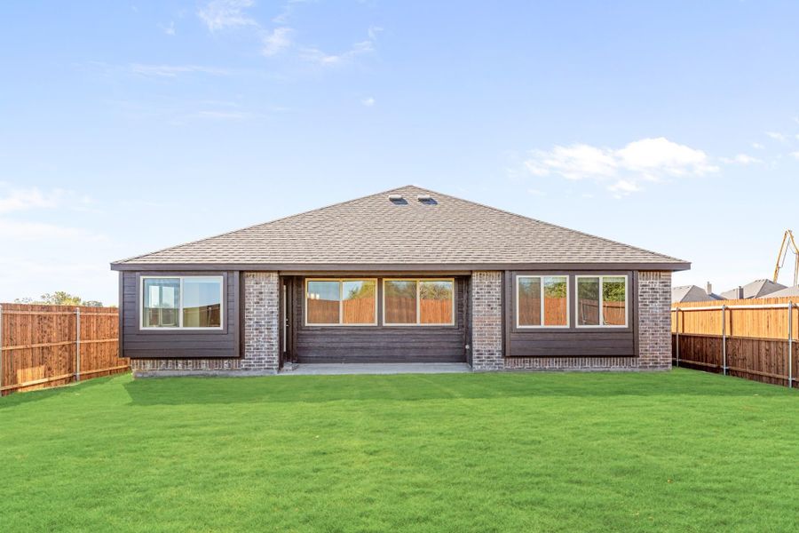 Rear view of single-story home with dark siding, brick accents, large windows, patio, and fenced backyard with green lawn