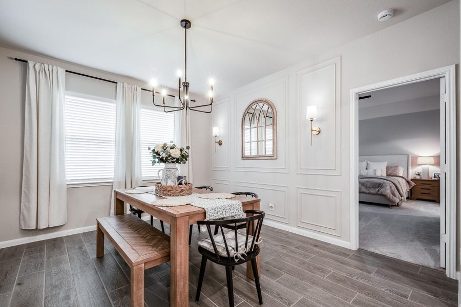 This photo showcases a bright dining area with a modern chandelier, wooden table, and cozy seating. The room features elegant wall paneling and a decorative mirror. Large windows with curtains provide ample natural light, and an adjoining bedroom is visible through an open doorway. This photo showcases a bright dining area with a modern chandelier, wooden table, and cozy seating. The room features elegant wall paneling and a decorative mirror. Large windows with curtains provide ample natural light, and an adjoining bedroom is visible through an open doorway.