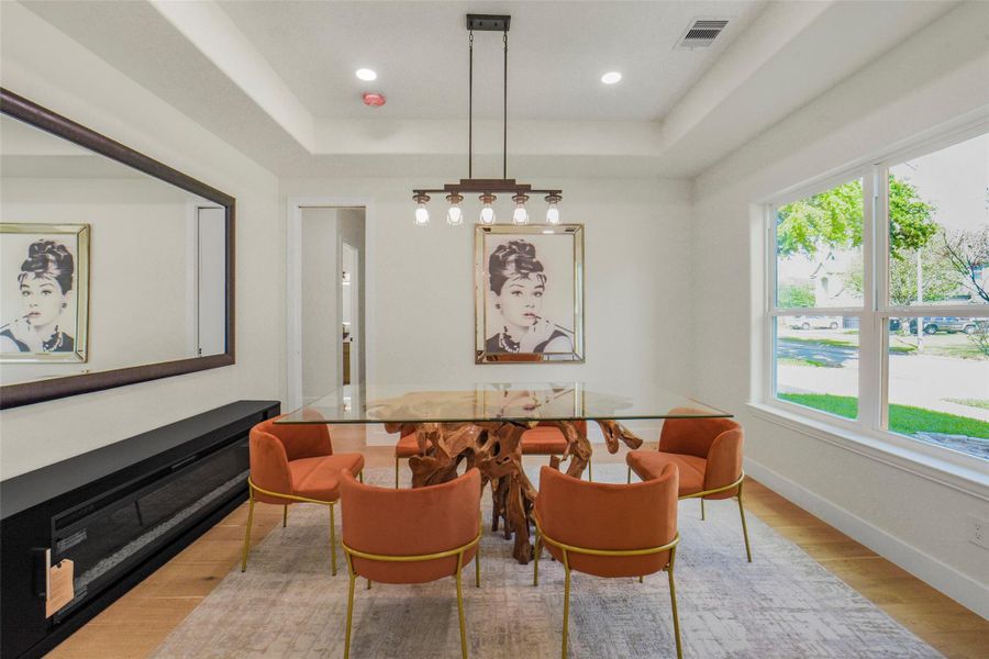 This dining room features a modern design with a large glass-top table and stylish orange chairs. The space is brightened by large windows and a contemporary light fixture. A wall mirror and elegant artwork add sophistication, while wood flooring provides warmth.