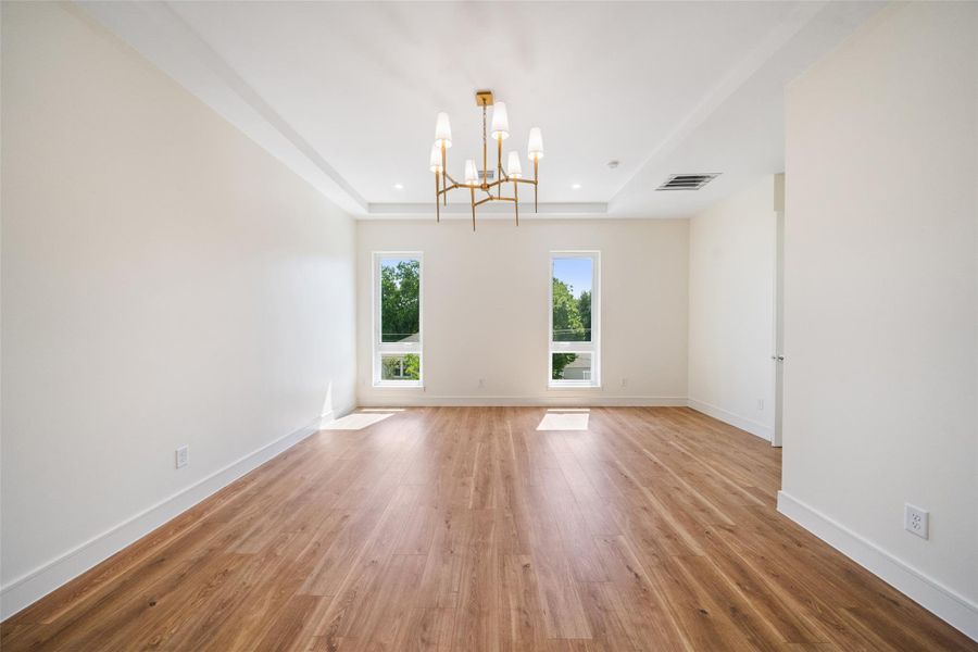 Primary bedroom features a spacious, light-filled area with two tall windows, wood flooring, and a modern chandelier. The neutral walls and recessed lighting provide a bright and airy feel.