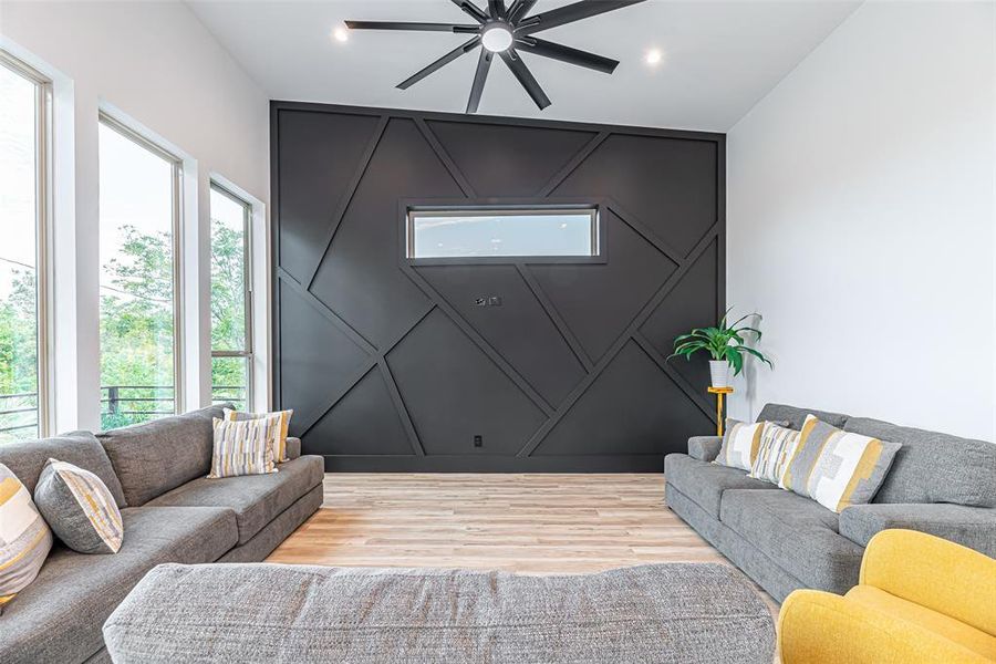 Modern living space with high ceilings, large windows, and a striking geometric accent wall. Features comfortable seating and a stylish ceiling fan.