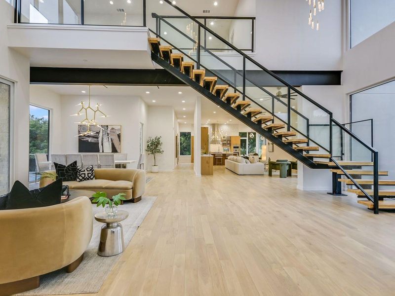 A grand floating staircase with sleek glass railings anchors this open-concept living space, showcasing light hardwood floors and high ceilings. A grand floating staircase with sleek glass railings anchors this open-concept living space, showcasing light hardwood floors and high ceilings.
