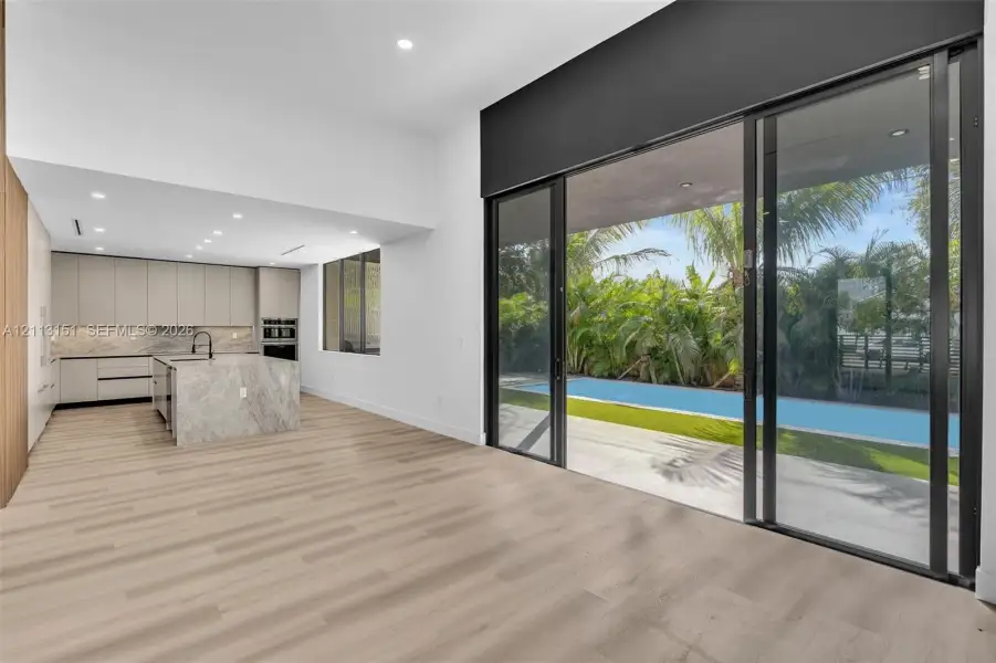 Interior space featuring light-toned wood-finish flooring, floor-to-ceiling sliding glass doors, kitchen island with stone-surface waterfall edge, flat-panel cabinetry, and integrated outdoor pool area Interior space featuring light-toned wood-finish flooring, floor-to-ceiling sliding glass doors, kitchen island with stone-surface waterfall edge, flat-panel cabinetry, and integrated outdoor pool area
