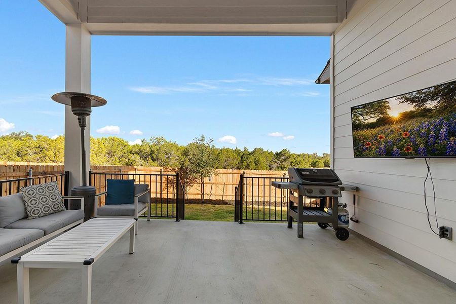 A spacious covered patio overlooks the fenced backyard and surrounding green trees under clear blue skies.