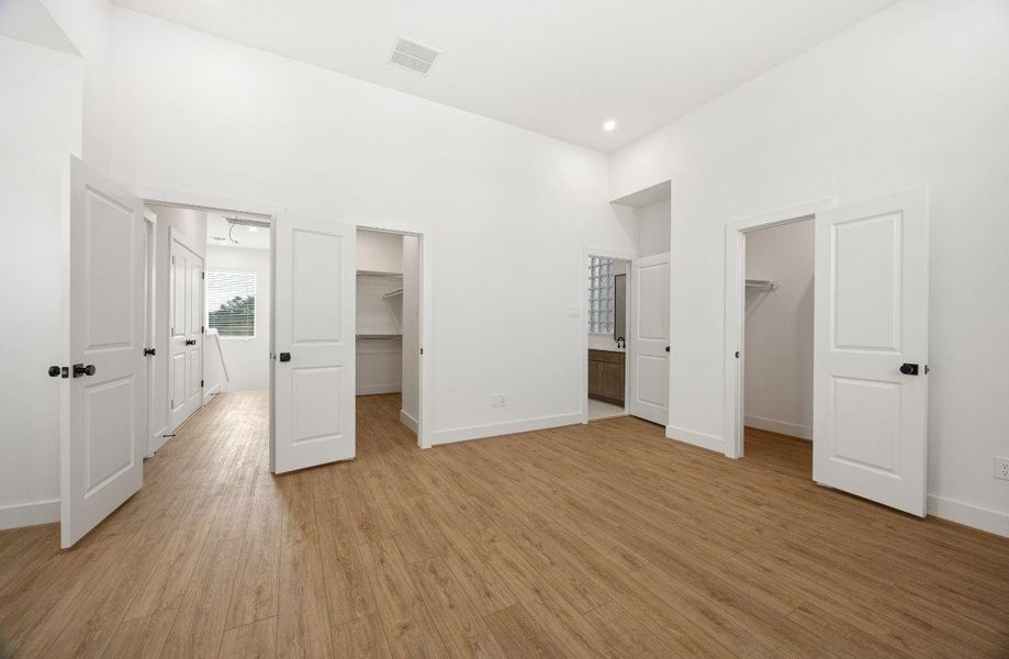Large bedroom suite with high ceilings, wood-look flooring, crisp white walls, and multiple access points to closets and bath areas. The open layout provides excellent flexibility, while the clean modern finishes, black hardware, and abundant storage create a polished and practical private retreat.
