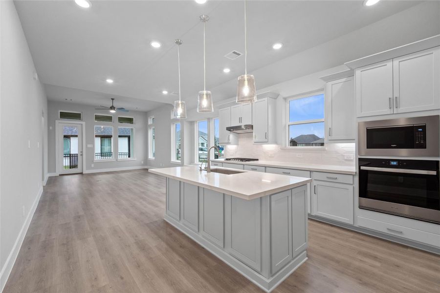 The open-concept gourmet kitchen is designed for both cooking and entertaining. It features elegant cabinetry, pristine quartz countertops, and a large kitchen island that doubles as a functional breakfast bar. The cooking area is a chef's dream with a sleek stainless steel gas cooktop, hood vent, and oven/microwave wall unit, all complemented by a chic tile backsplash.