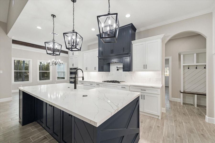 View from kitchen toward garage entry hall on the right.  Arched opening shows book/nook bench with coat hooks just inside the garage entry.   Far left is the beam ceiling and wall of windows in the dining room.  Kitchen island has pop-up charger and electric plugs.
