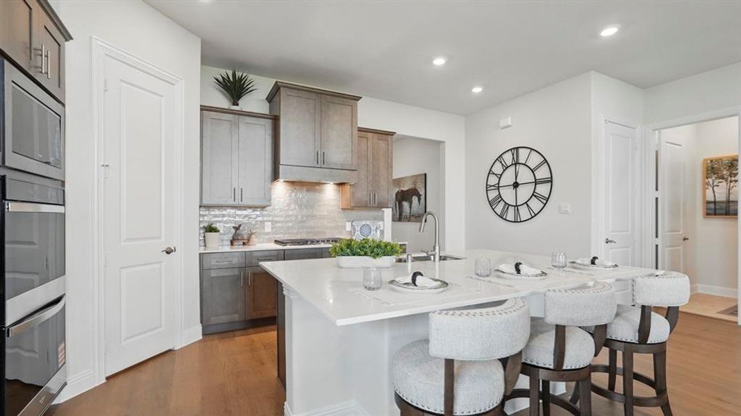 Modern kitchen featuring an expansive island with a white countertop and seating, stainless steel double ovens, gray wood-finish cabinetry, a subway tile backsplash, and recessed lighting