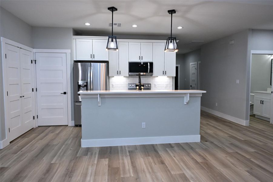 Kitchen with white cabinetry, a kitchen island with sink, stainless steel appliances, hanging light fixtures, and decorative backsplash