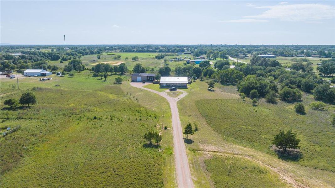 Expansive rural property featuring a main building with a lighter roof and a darker facade, a separate outbuilding, a gravel driveway, and mature trees dotting the landscape Expansive rural property featuring a main building with a lighter roof and a darker facade, a separate outbuilding, a gravel driveway, and mature trees dotting the landscape