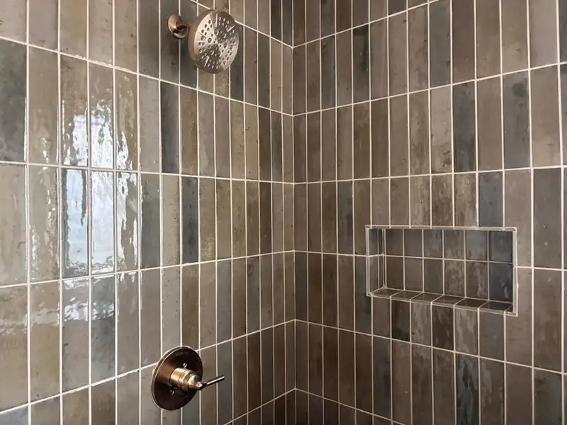 Shower stall featuring vertical subway tiles in varied earth tones, a recessed niche for storage, a brushed nickel shower head, and a matching shower control handle
