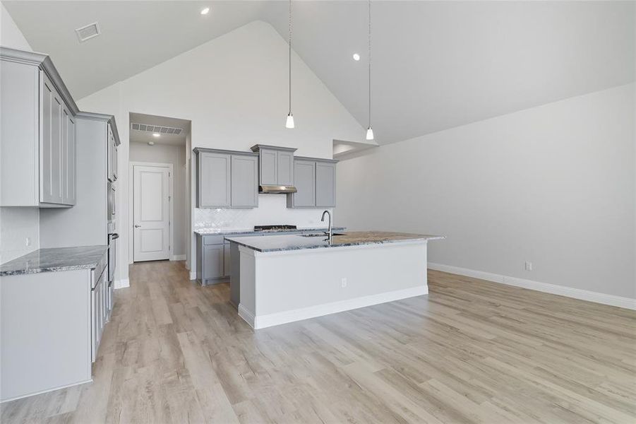 Kitchen featuring gray cabinetry, a high ceiling, a center island with sink, light stone countertops, and light wood-style flooring