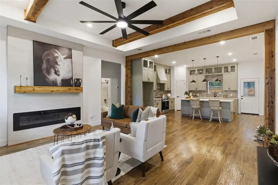 Open concept living area featuring a linear fireplace with wood mantel, wood-finish flooring, exposed wood beams, and a ceiling fan