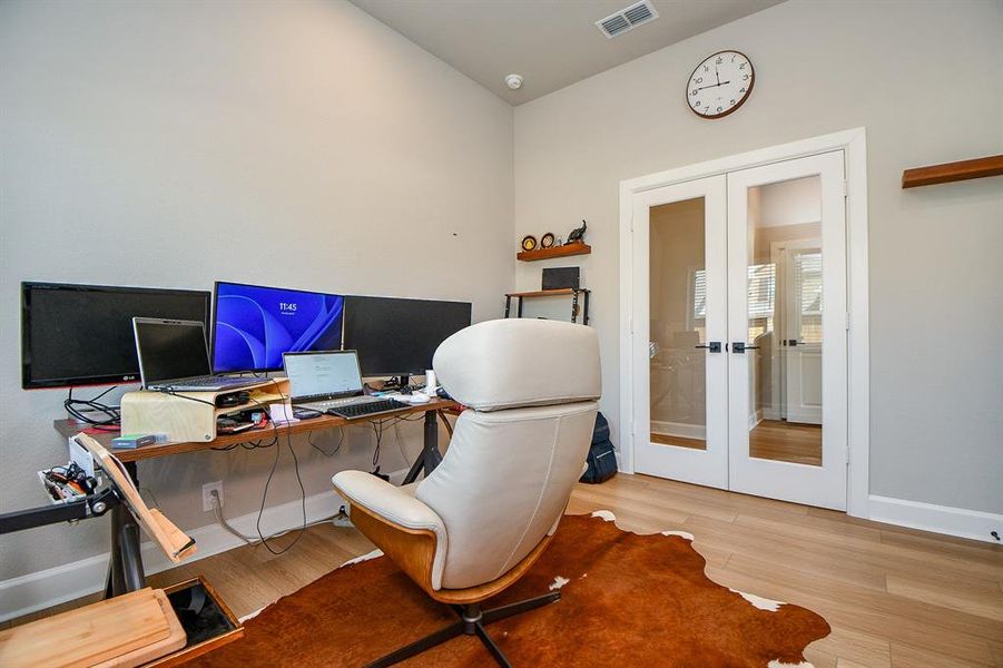 This home office features a modern setup with multiple monitors on a spacious desk, a comfortable chair, and light wood flooring. It includes glass double doors for a bright, open feel, and minimalist decor with a wall clock and shelving.