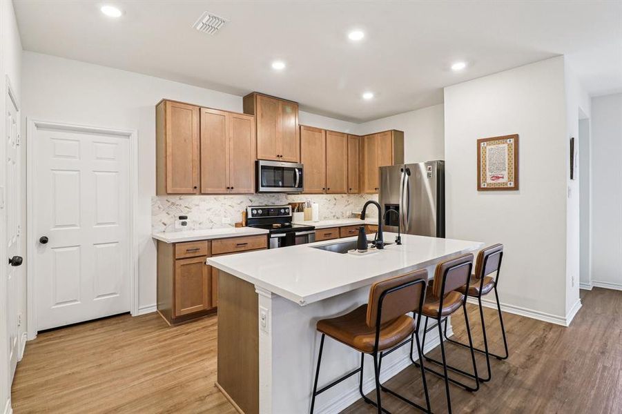 Kitchen with a breakfast bar, decorative backsplash, a kitchen island with sink, stainless steel appliances, and light wood-style floors Kitchen with a breakfast bar, decorative backsplash, a kitchen island with sink, stainless steel appliances, and light wood-style floors